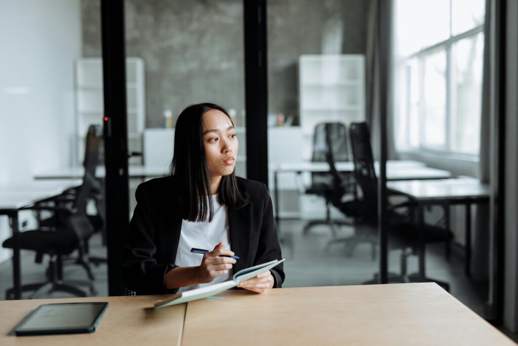 a girl sitting at a desk in an office, holding a book and a pen and looking pensively into the distance